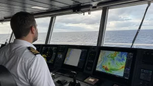 Gemi hava raporları değerlendiren vardiya zabiti köprüüstünde deniz hava raporu, swell ve rota planlama bilgilerini inceliyor.